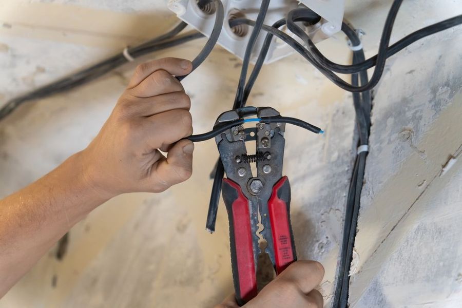 A person uses wire strippers on a black electrical cable inside a wall.