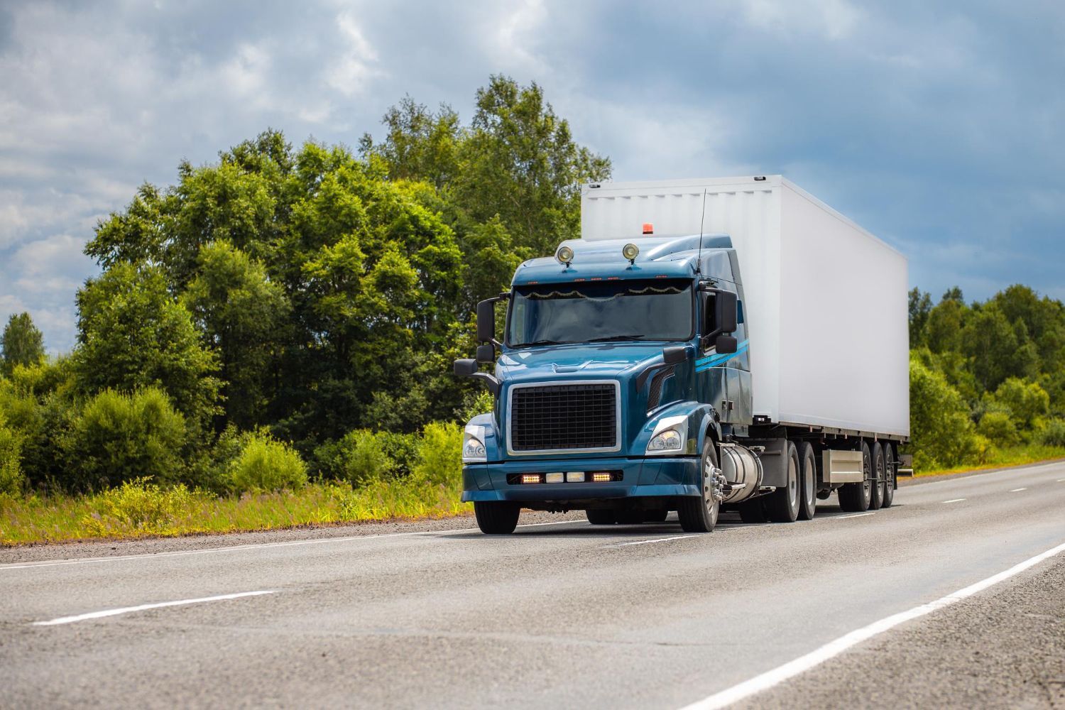 Blue semi-truck driving on a road with green trees in the background under a cloudy sky.