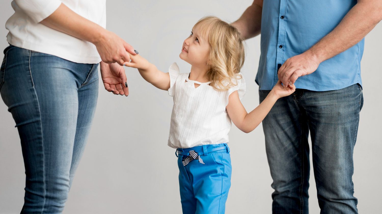 A young child holds hands with two adults. The child wears a white top and blue pants, looking up.
