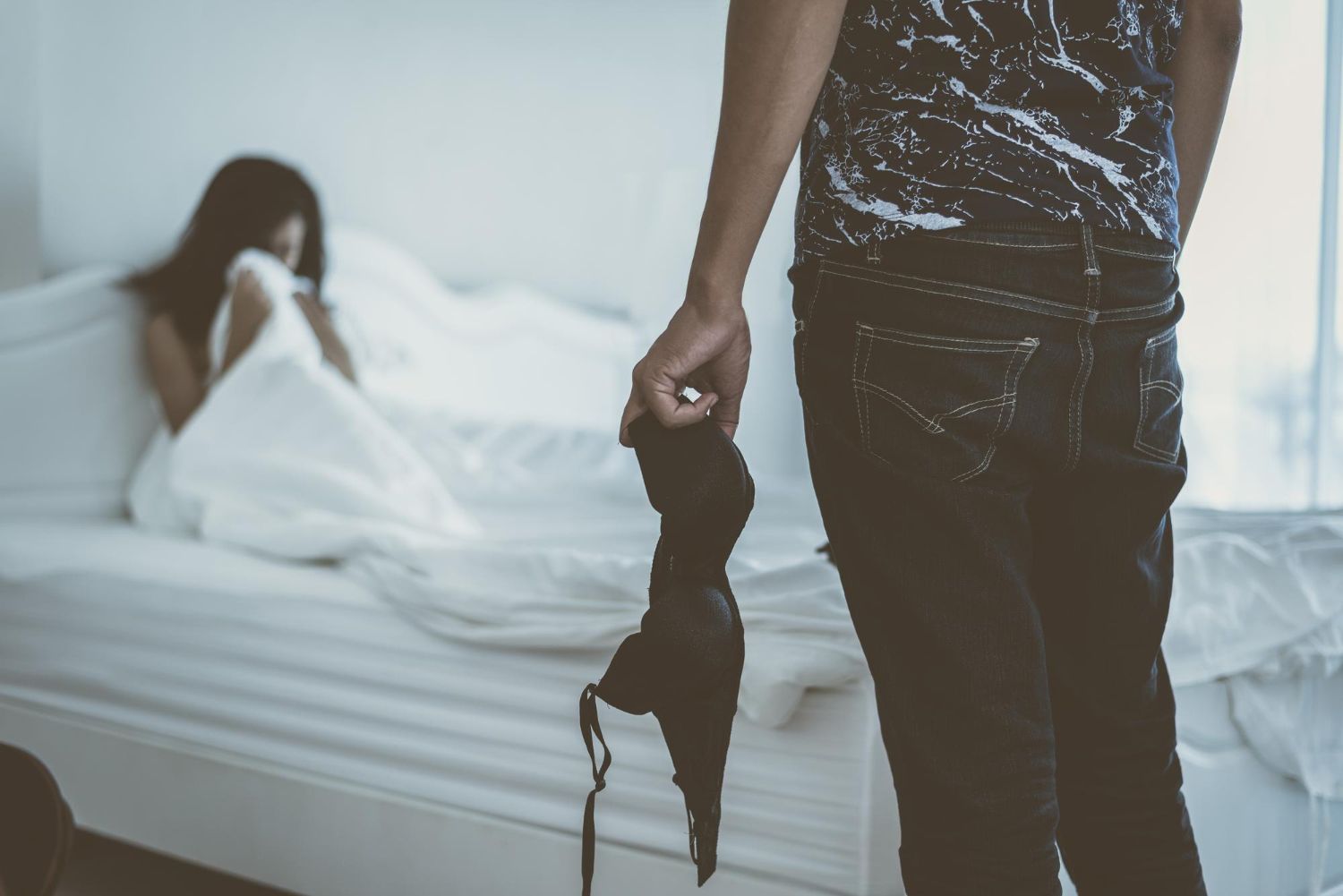 Person holding a black bra, woman on a bed covered by a white blanket.