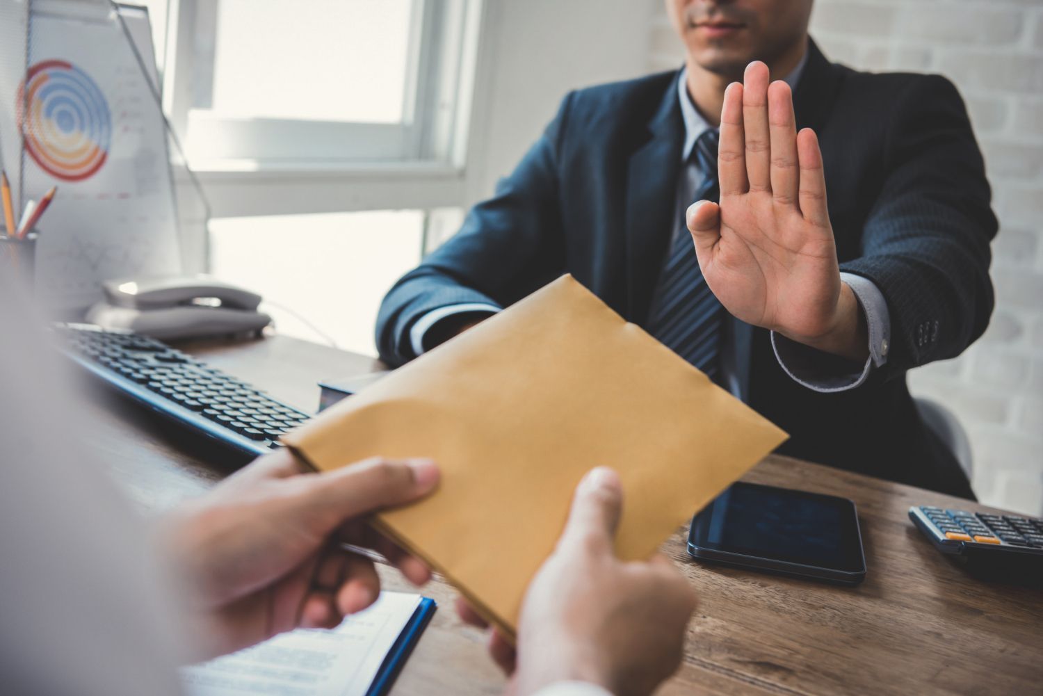 Man rejecting bribe: person in suit blocks hand, the other offers a brown envelope at a desk.