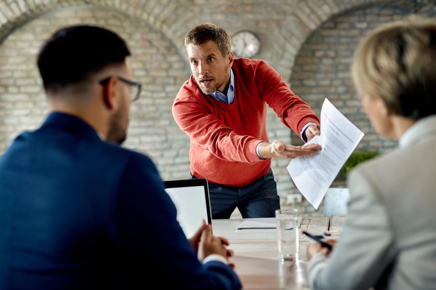 Man in red sweater gestures angrily, holding papers in meeting with two colleagues.