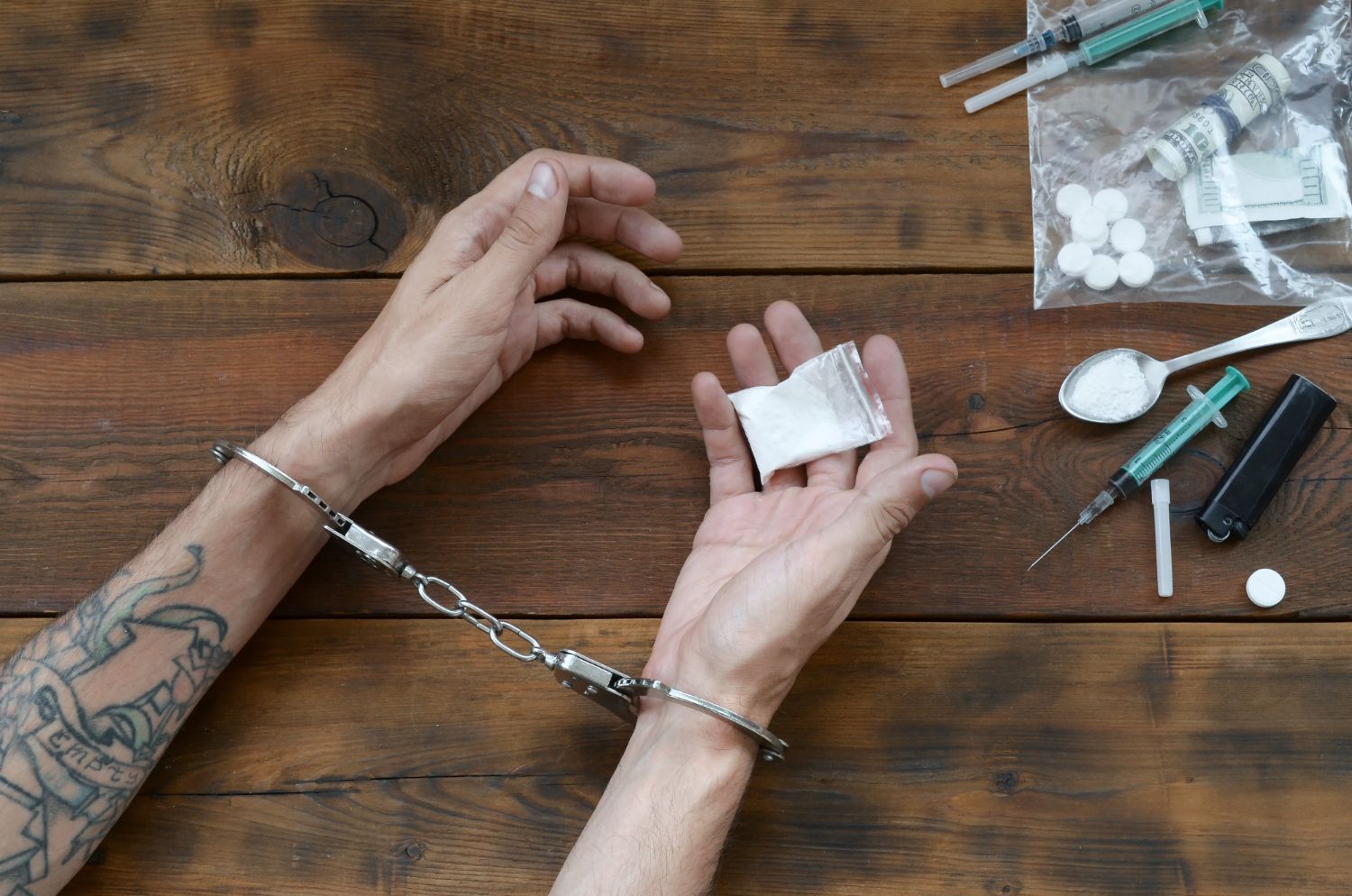 Hands in handcuffs holding a bag of white powder, with drug paraphernalia on a wooden table.