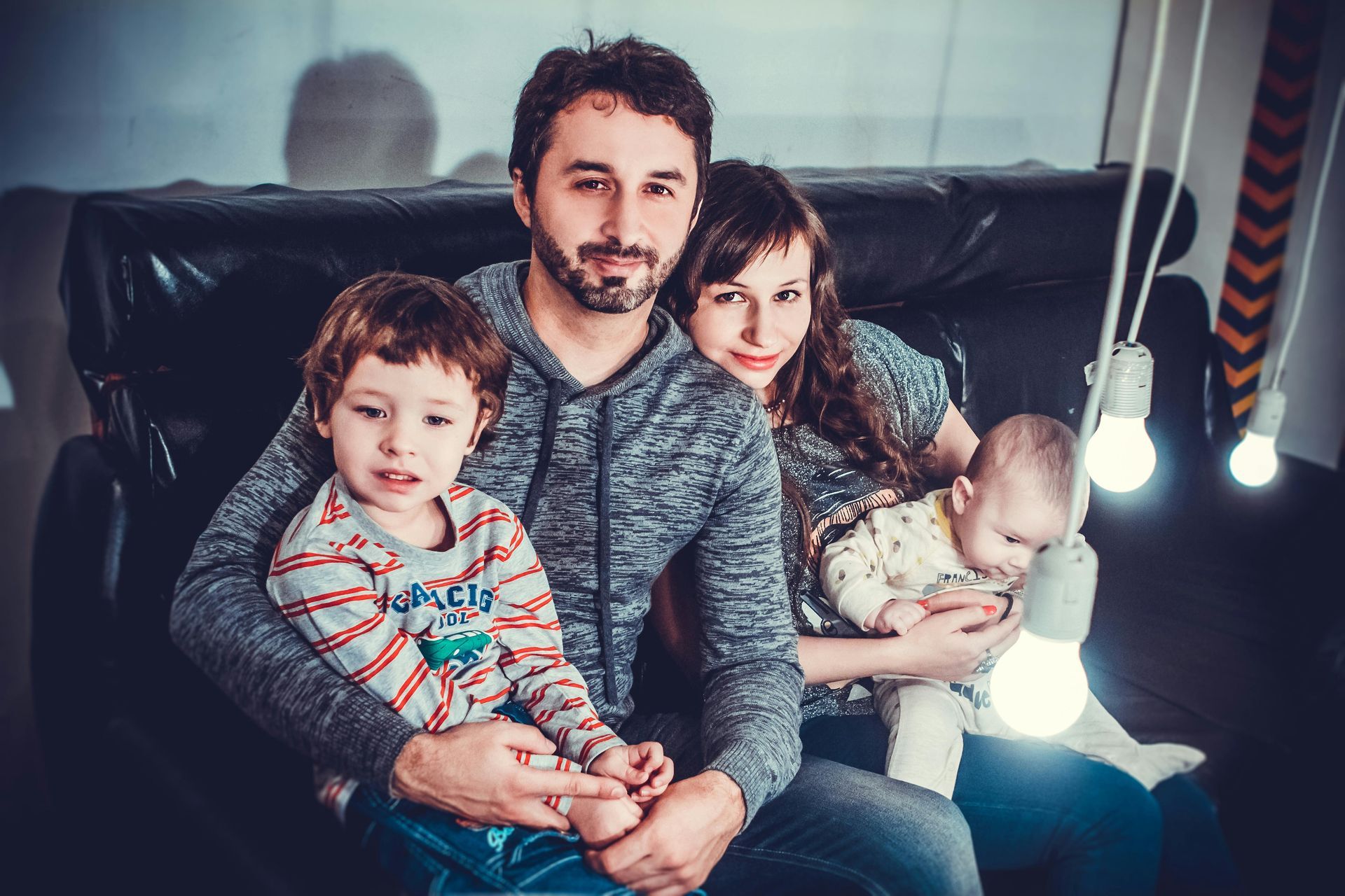 Family of four sitting on a black couch, illuminated by hanging light bulbs.