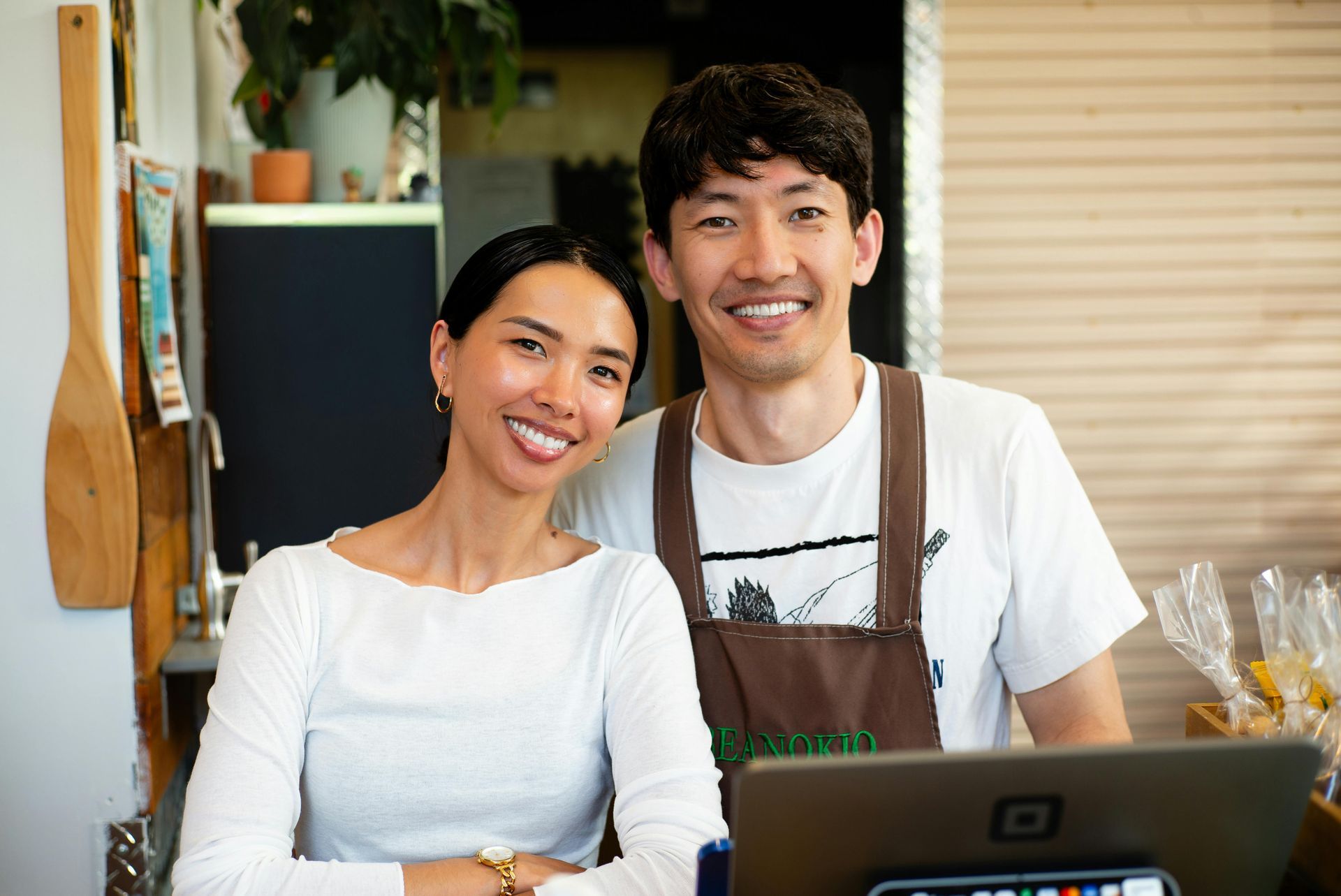 Two smiling people behind a counter with a laptop. One wears a white shirt, the other an apron.