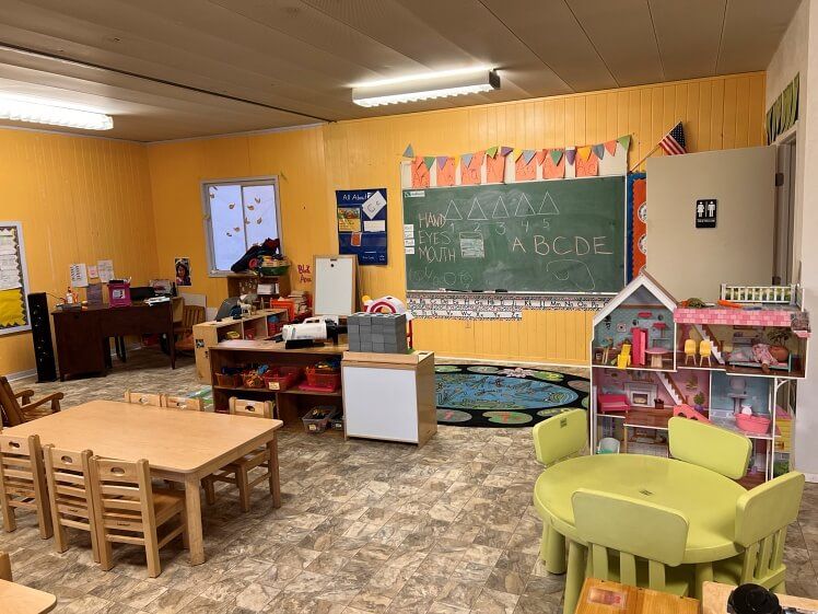 A classroom with tables , chairs , a doll house and a blackboard.