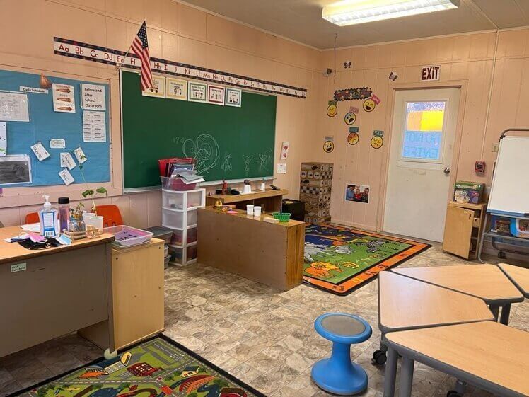 A classroom with tables , chairs , a rug and a green board.