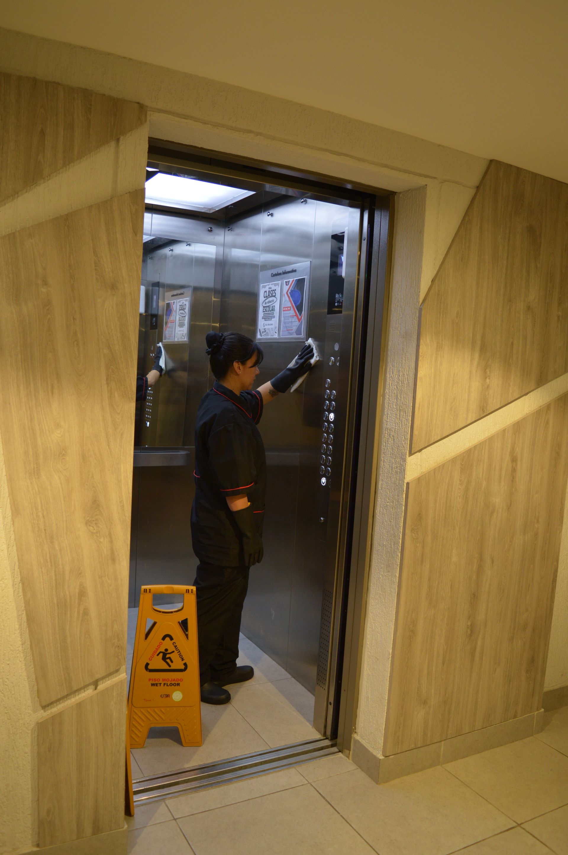 Una mujer está limpiando la puerta de un ascensor junto a una señal de precaución.
