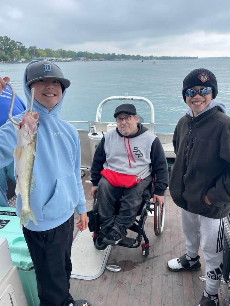 A man in a wheelchair is fishing with two boys on a boat.