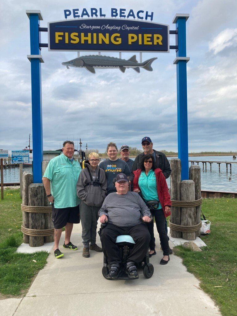 Group poses at Pearl Beach Fishing Pier sign, cloudy sky, pier in background.