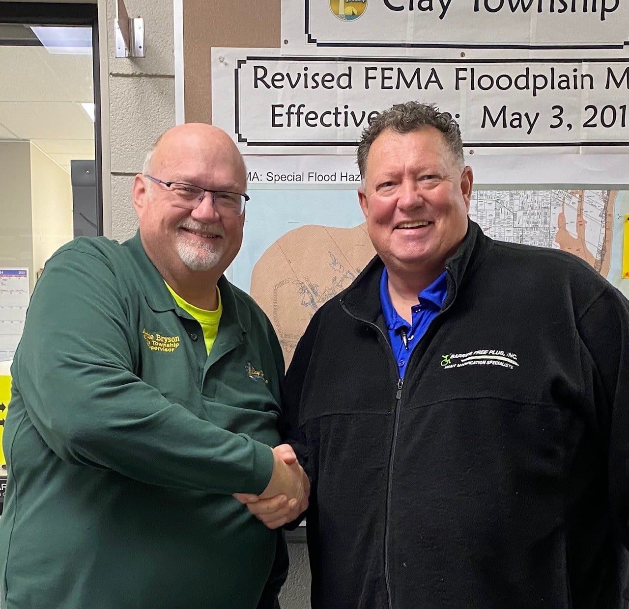 Two men shaking hands in front of a FEMA floodplain map. One wears green, the other black.
