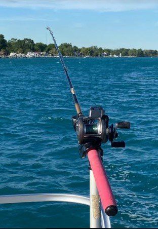 Fishing rod with reel mounted on a boat railing over blue water, bright day.