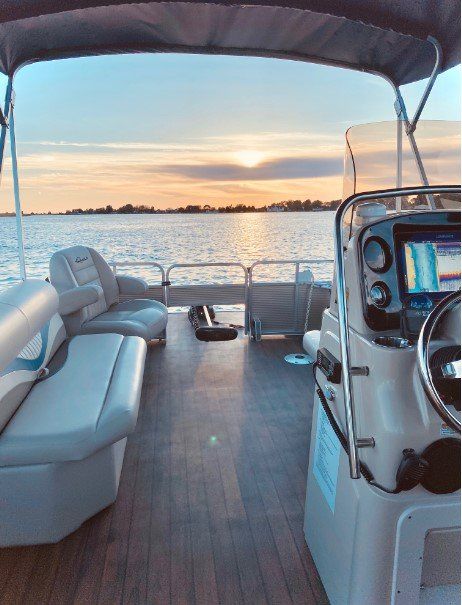 Interior of a pontoon boat overlooking a calm lake at sunset; wooden deck, white and gray seating.