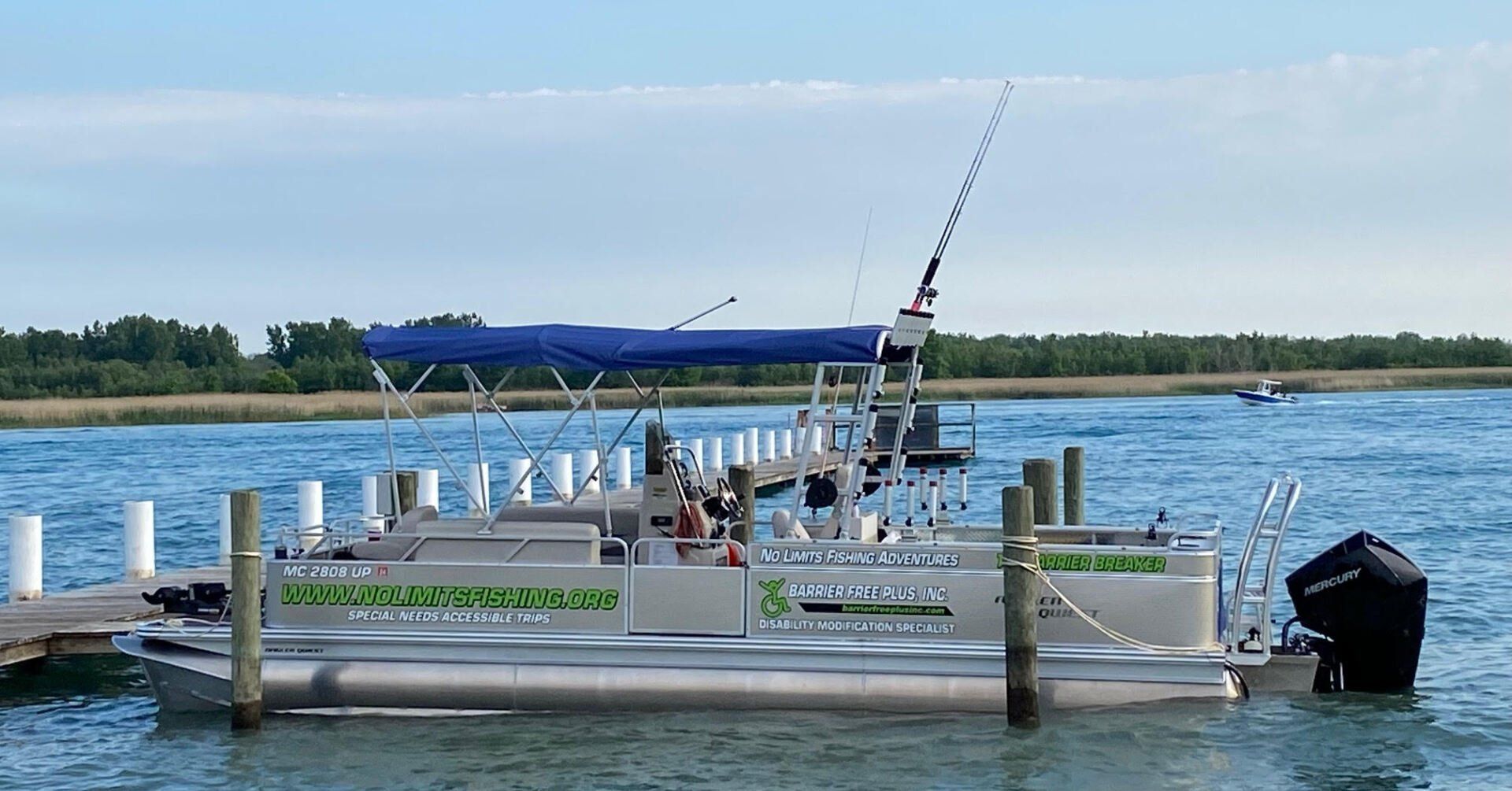 Pontoon boat docked at a pier, fishing rods visible. Blue canopy, water, and trees in background.