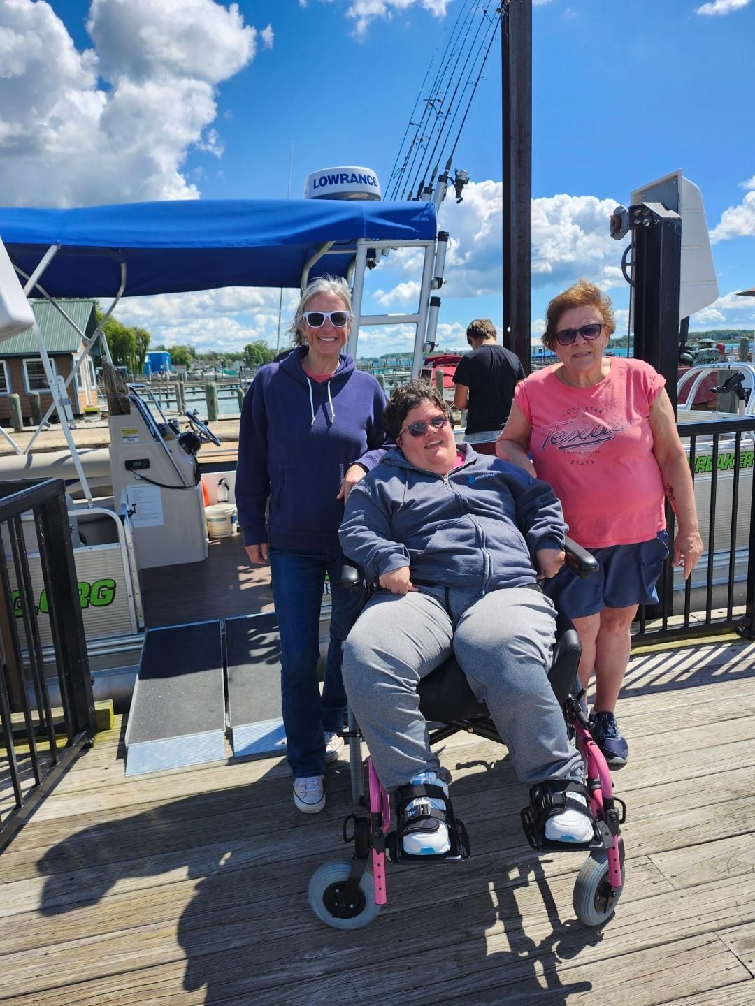 Three people on a dock; one in a wheelchair, two standing. Blue boat in background under a sunny sky.