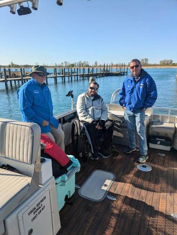 Three people on a boat near a dock; one in a wheelchair. Blue jackets, sunny day.