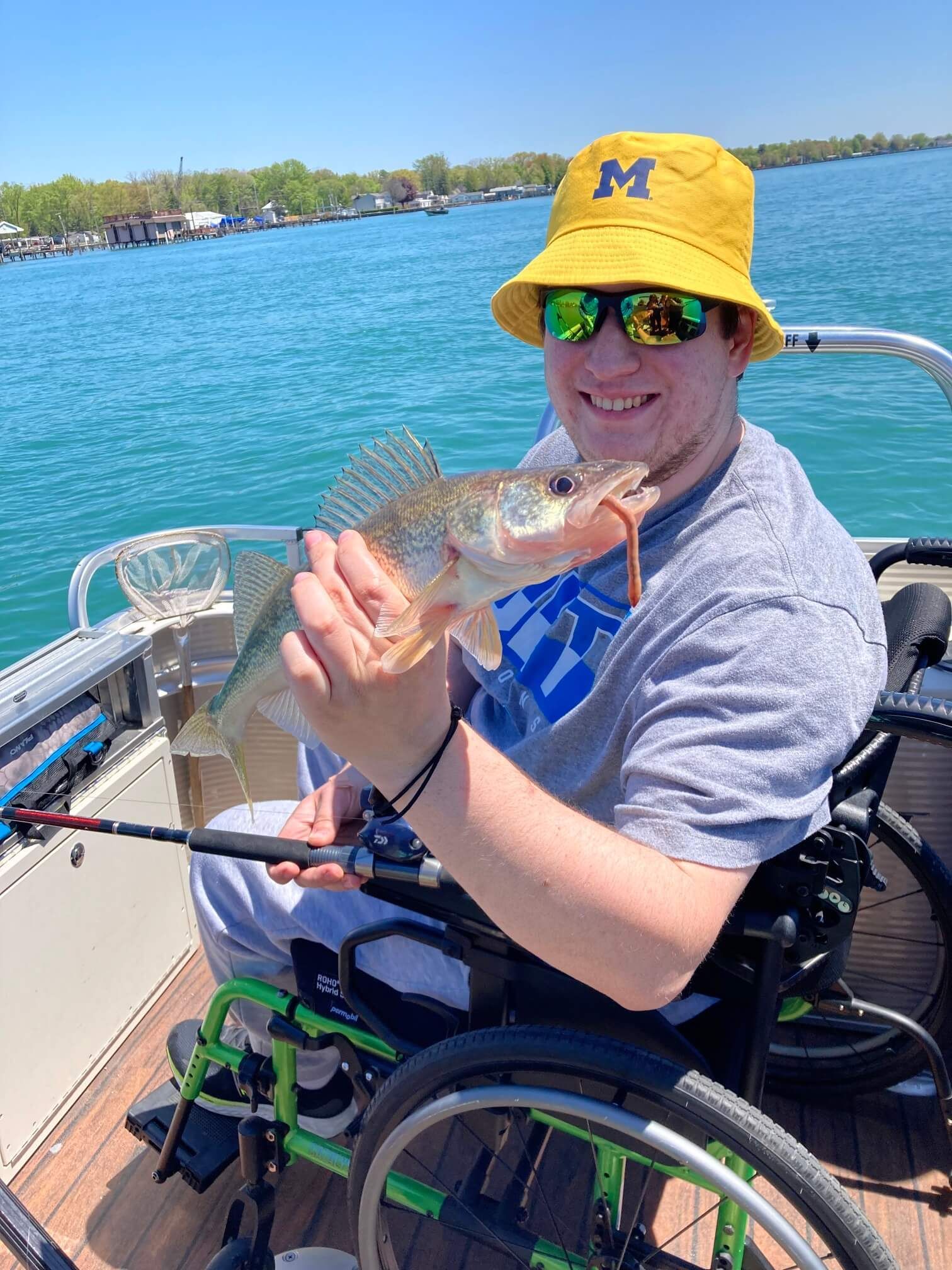 Man in wheelchair on a boat, holding a fish, wearing a yellow hat and sunglasses. Lake background.