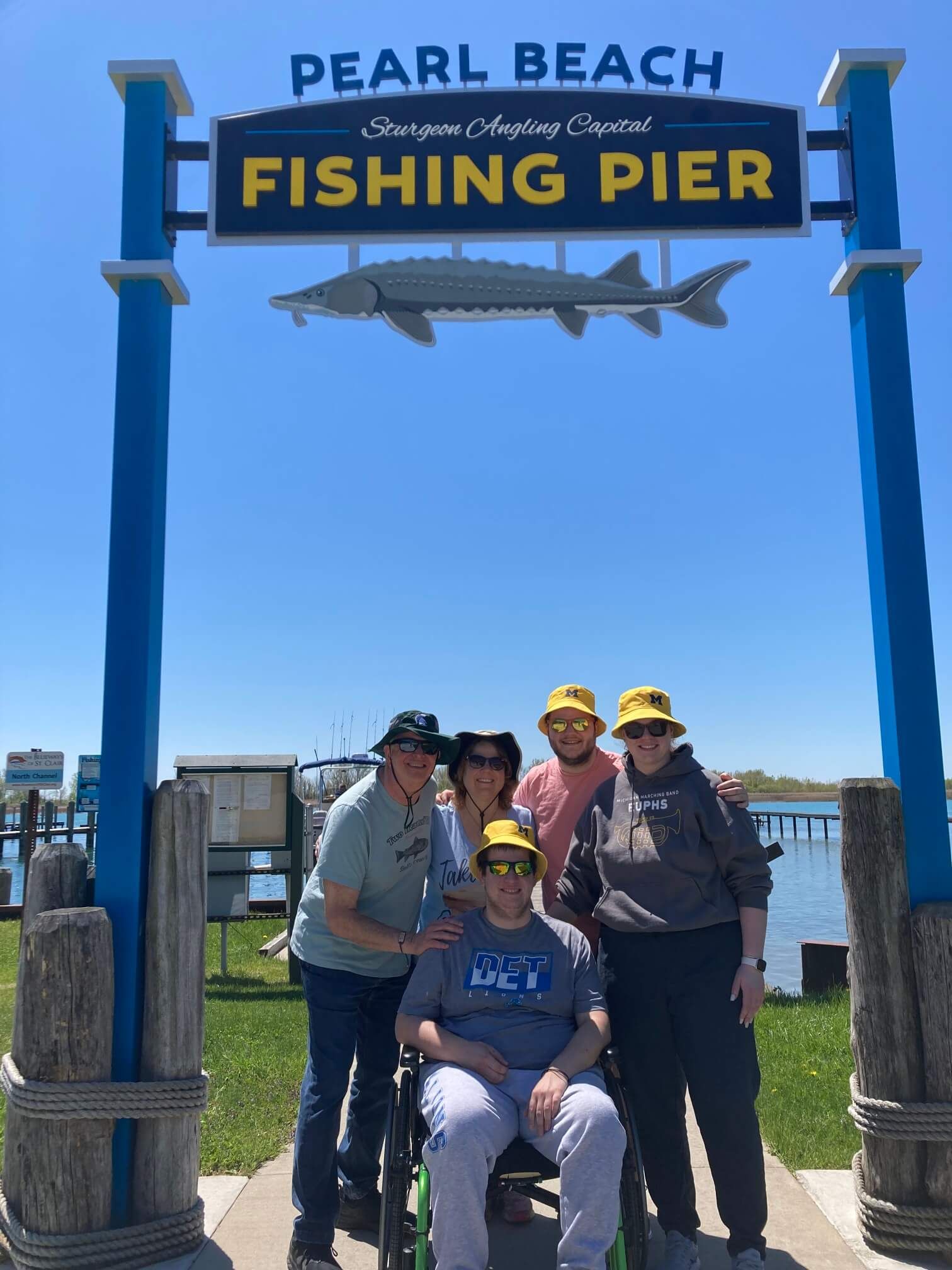 People pose at Pearl Beach Fishing Pier sign, person in wheelchair in front. Blue sky.