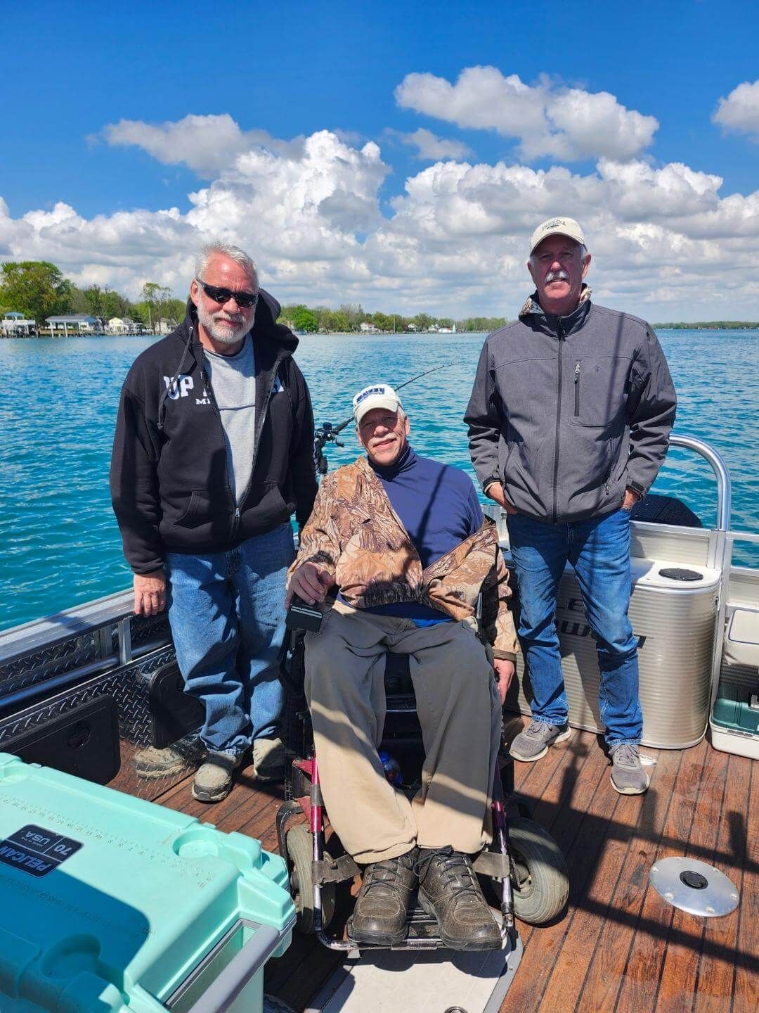 Three men on a boat, one in a wheelchair, on a sunny day. Blue water, cloudy sky, and shoreline in the background.