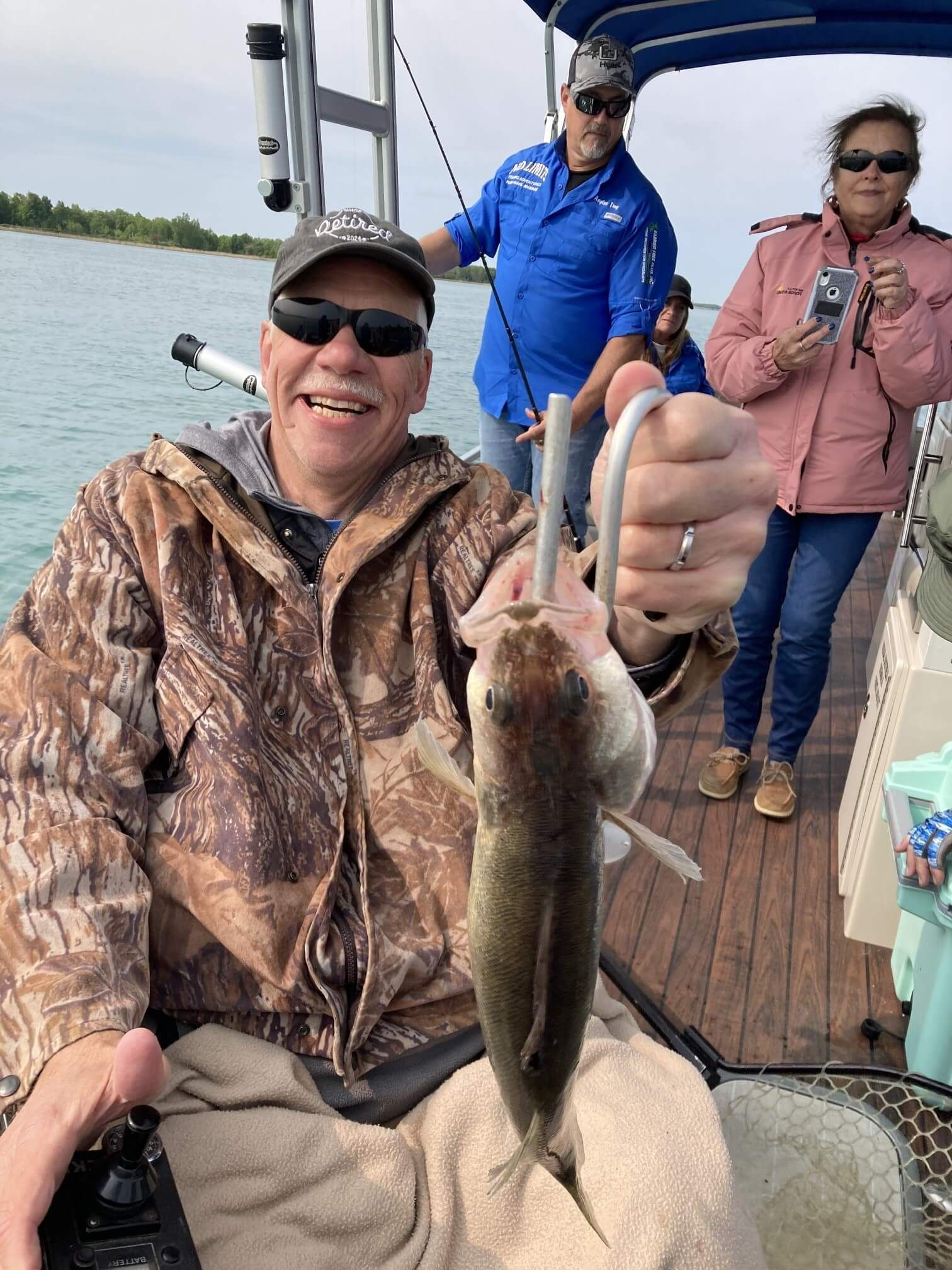 Man in camo holds fish on a boat; others fish. Blue water, overcast day.