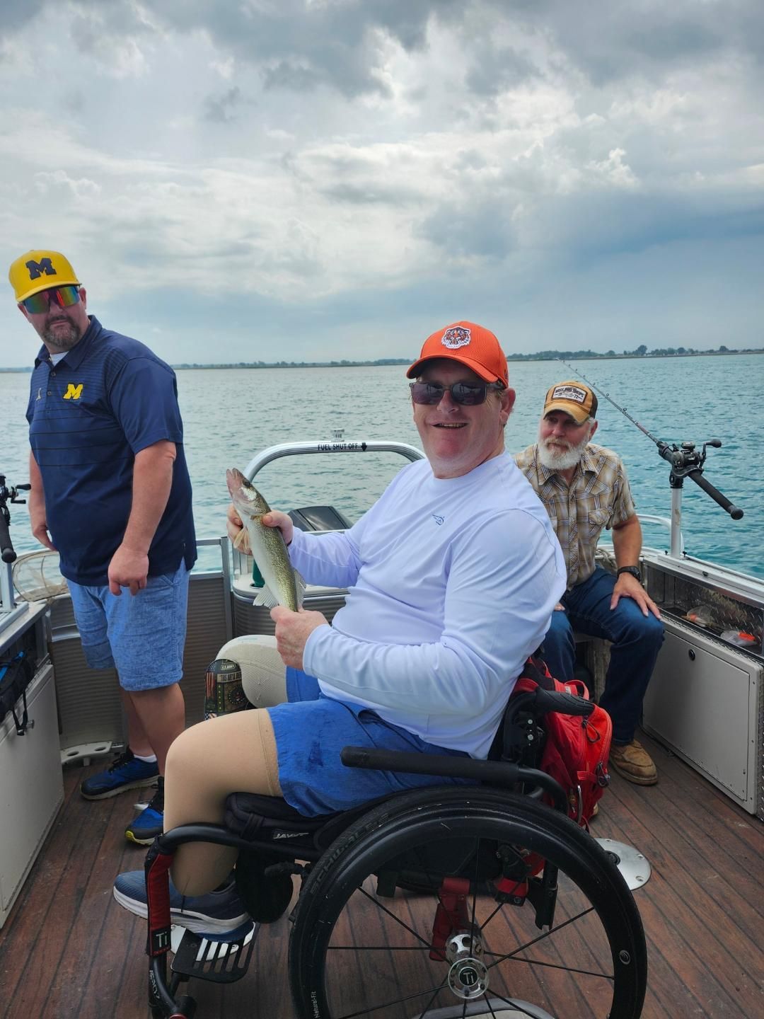 Man in wheelchair holding a fish on a boat; two other men watch in cloudy water.