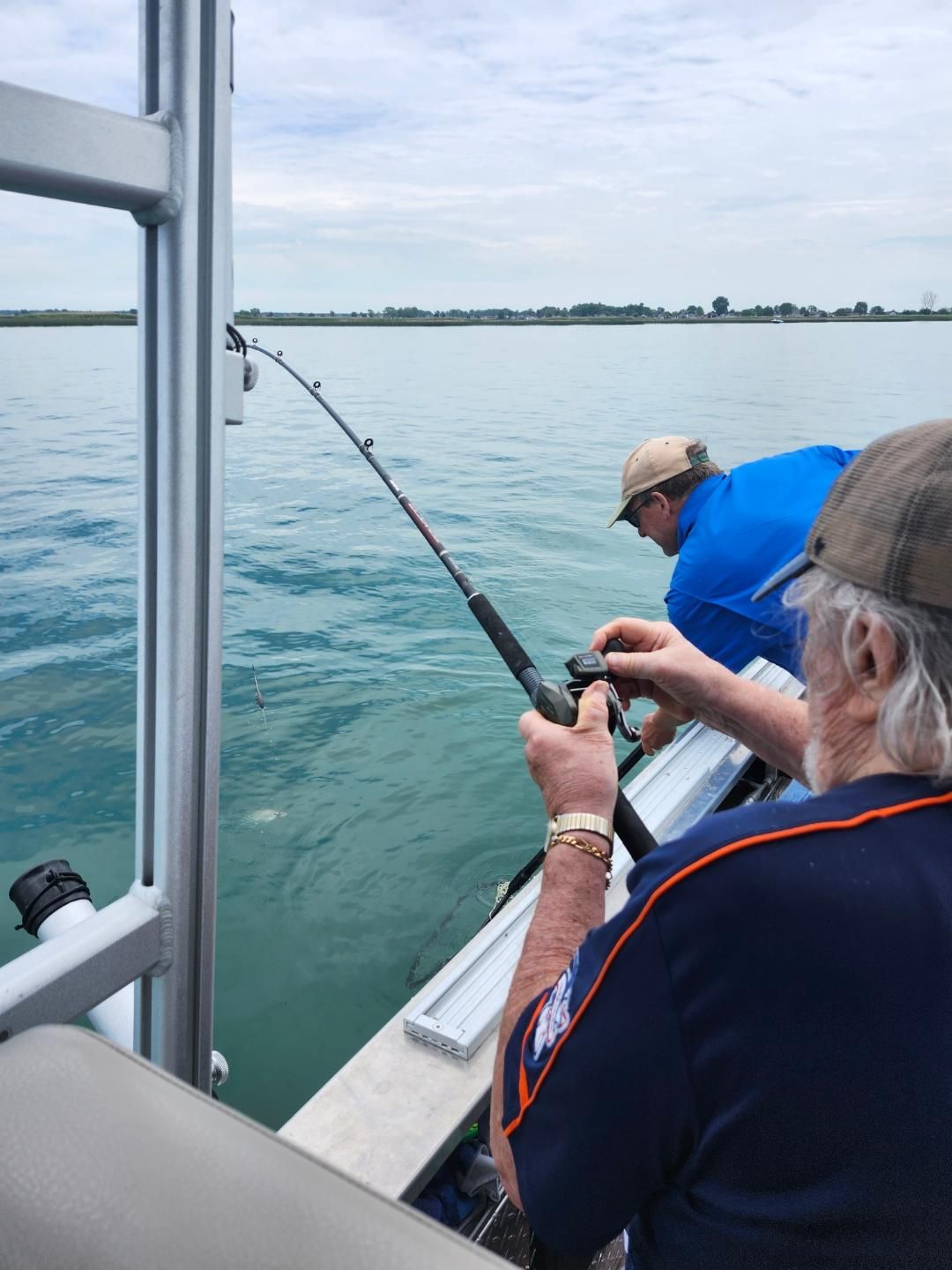 Two men fishing from a boat on a body of water. One reels in a rod, the other watches.