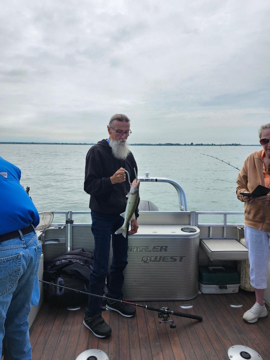 Man on a boat holding a fish. Cloudy sky, water in the background. Fishing poles visible.