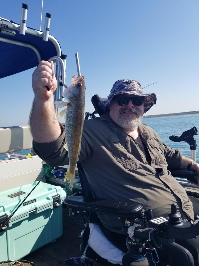 Man in a wheelchair holding up a fish on a boat. Sunglasses, hat, blue water, sunny day.