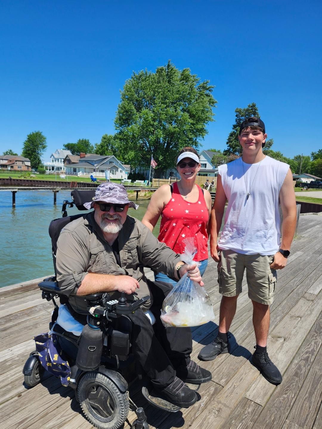 Three people pose on a wooden pier by a body of water. A person in a wheelchair holds a bag, smiling. The others stand nearby. Bright, sunny day.