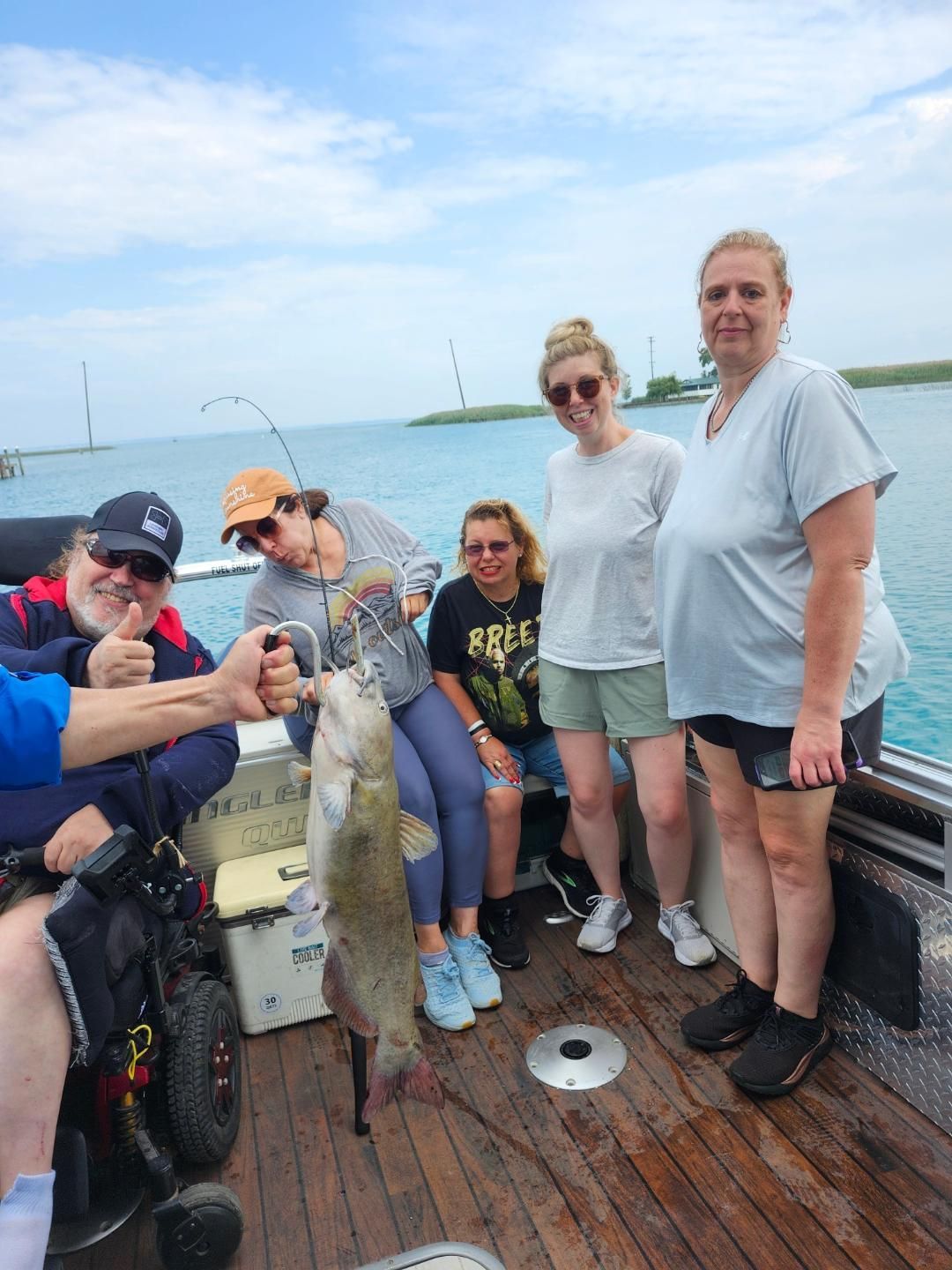 Group of people on a boat with a caught fish. Man in wheelchair gives thumbs up. lake in the background.