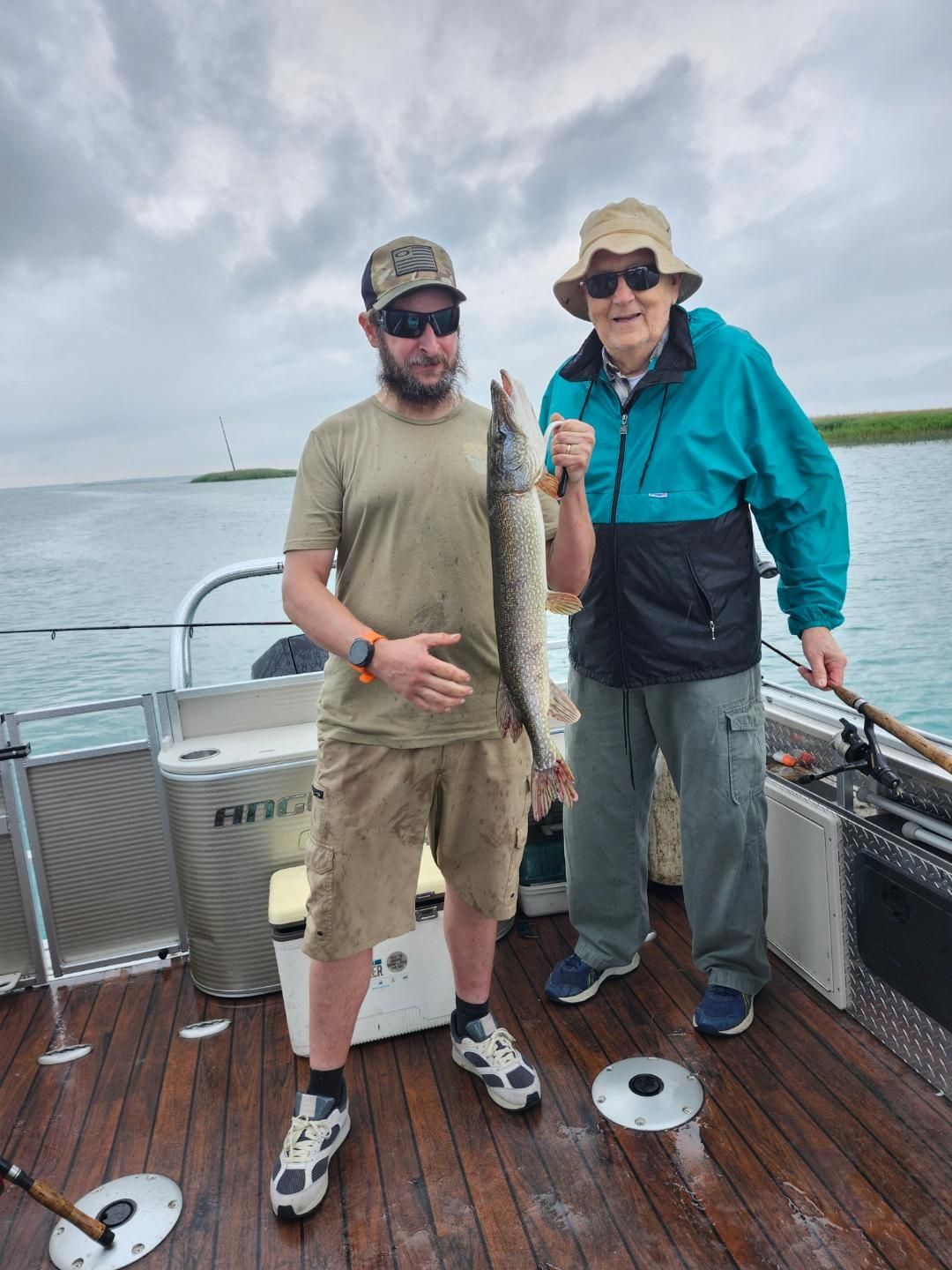 Two men on a boat, one holding a fish. Cloudy sky, water background.