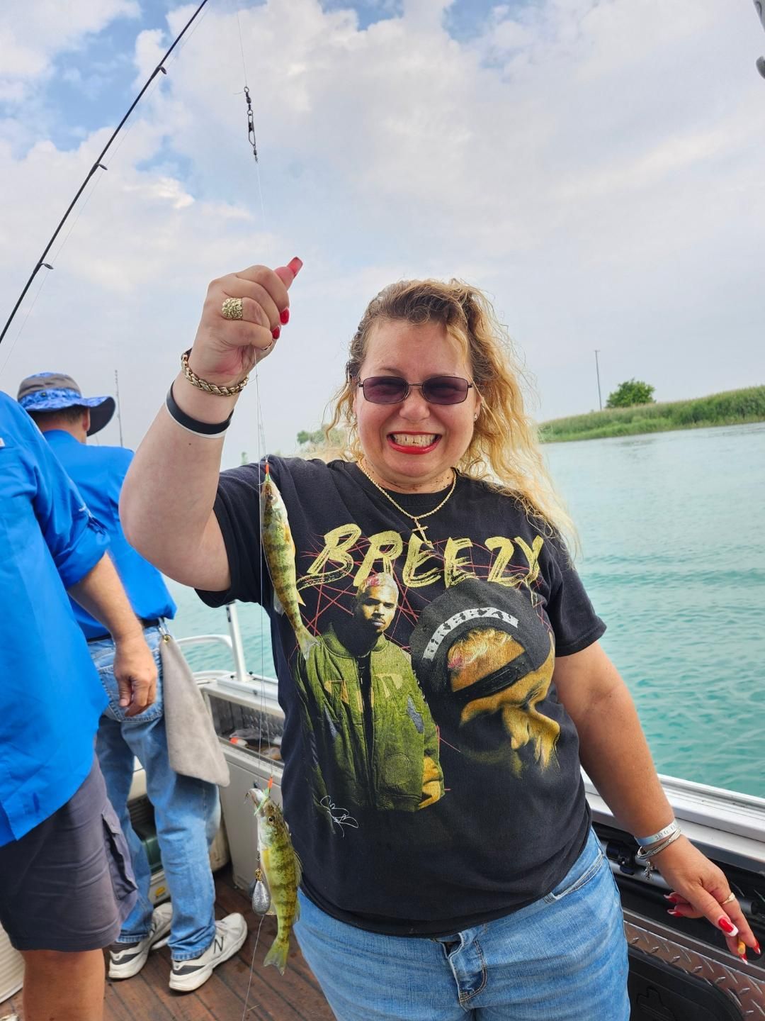 Woman on boat holding up fish she caught; smiling, wearing sunglasses and a black tee shirt.