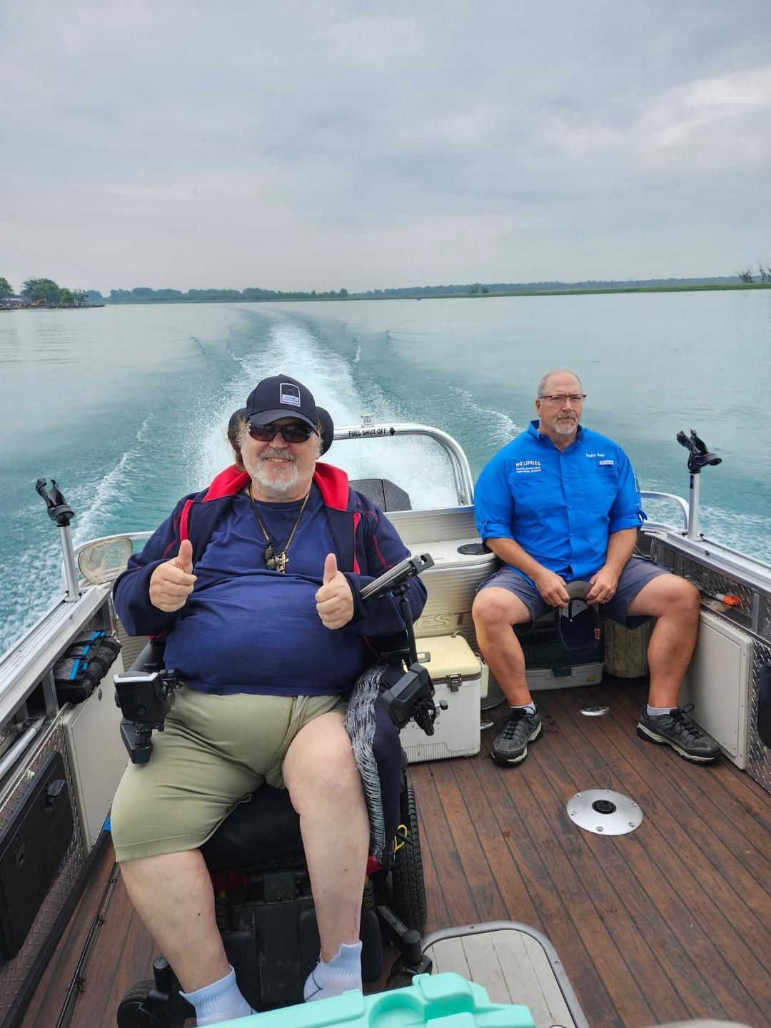 Two men on a boat, one in a wheelchair, thumbs up. The boat is on a body of water under an overcast sky.