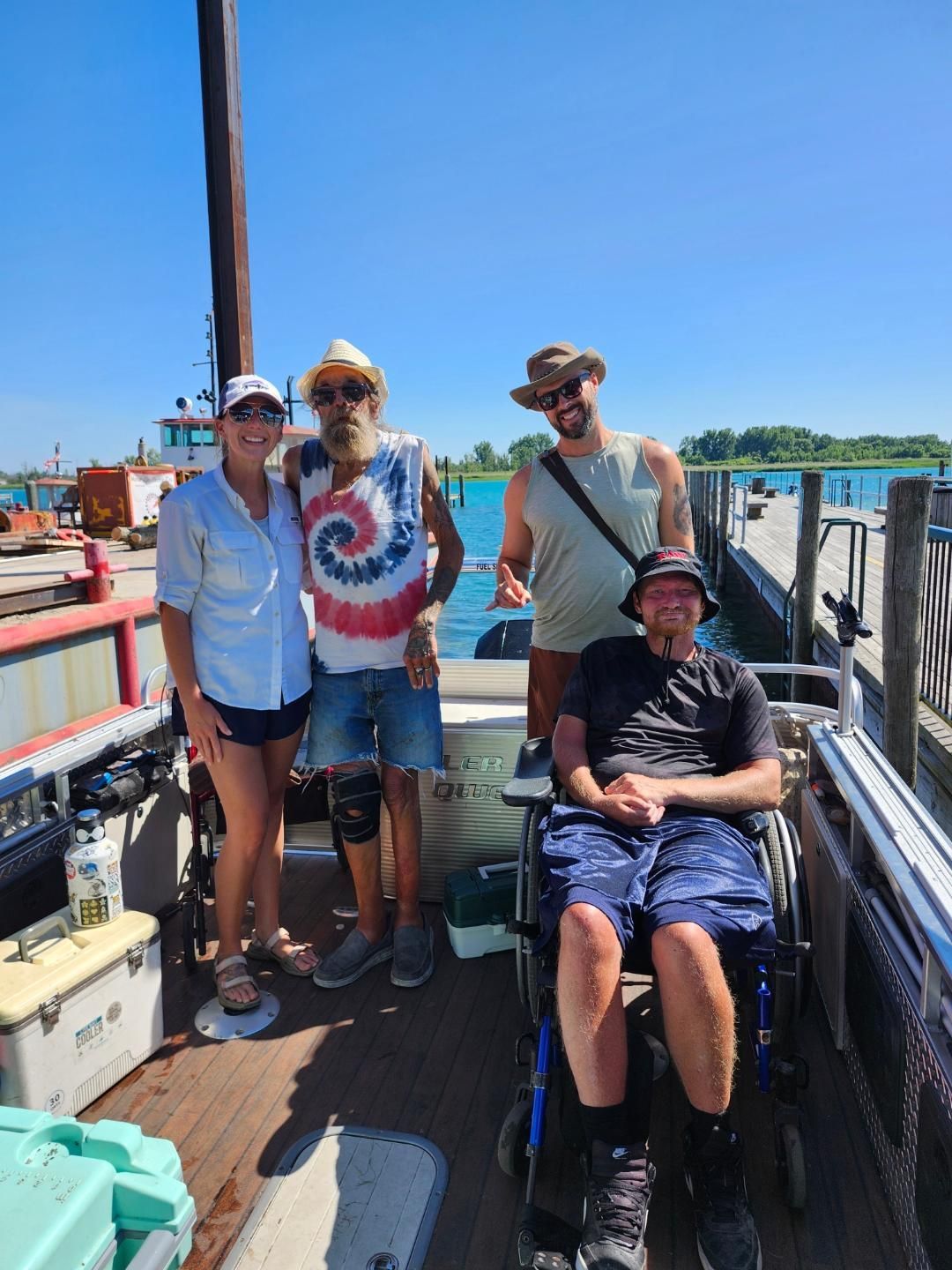 Four people pose on a boat dock, one in a wheelchair. Sunny day, blue water and sky.