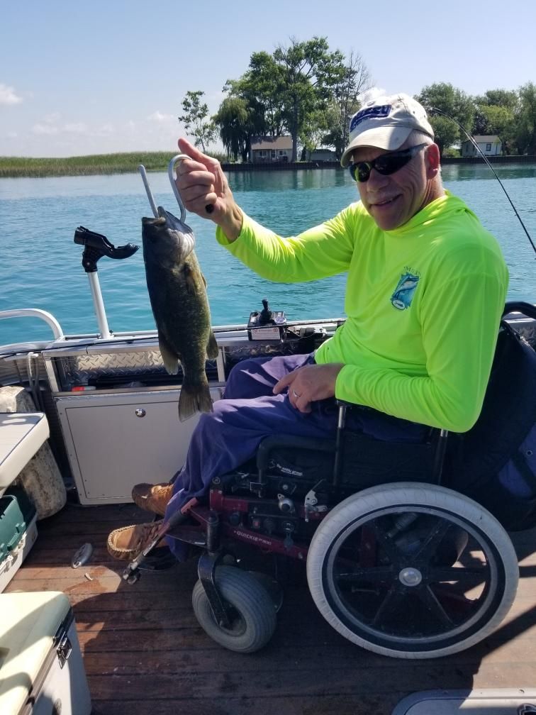 Man in wheelchair on a boat holding a fish he caught. Blue water, sunny day.