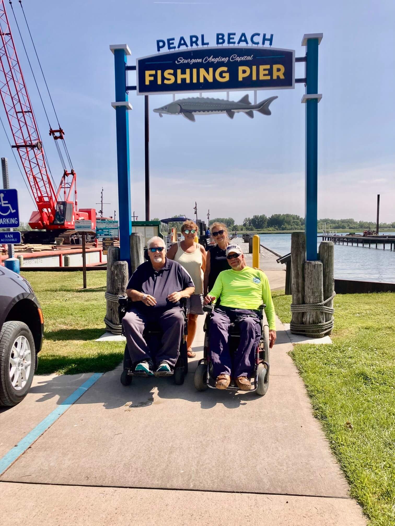 Group of people, including two in wheelchairs, by Pearl Beach Fishing Pier sign, sunny day.