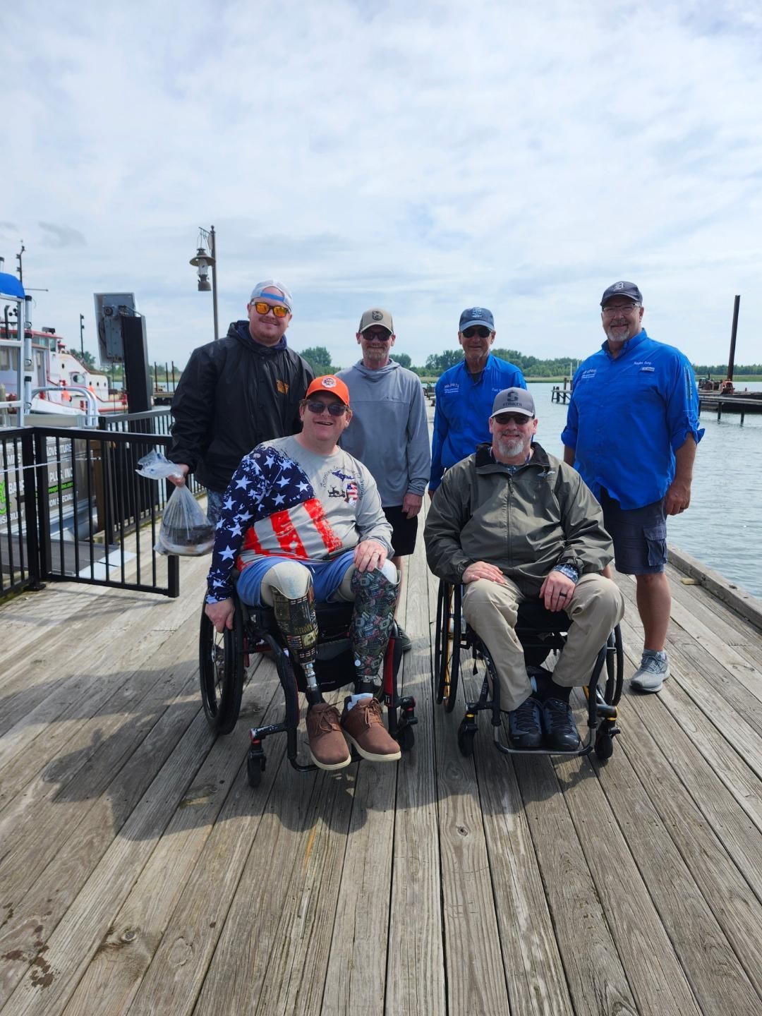Group of men on a wooden dock; two in wheelchairs. Cloudy sky, water, and boats in the background.