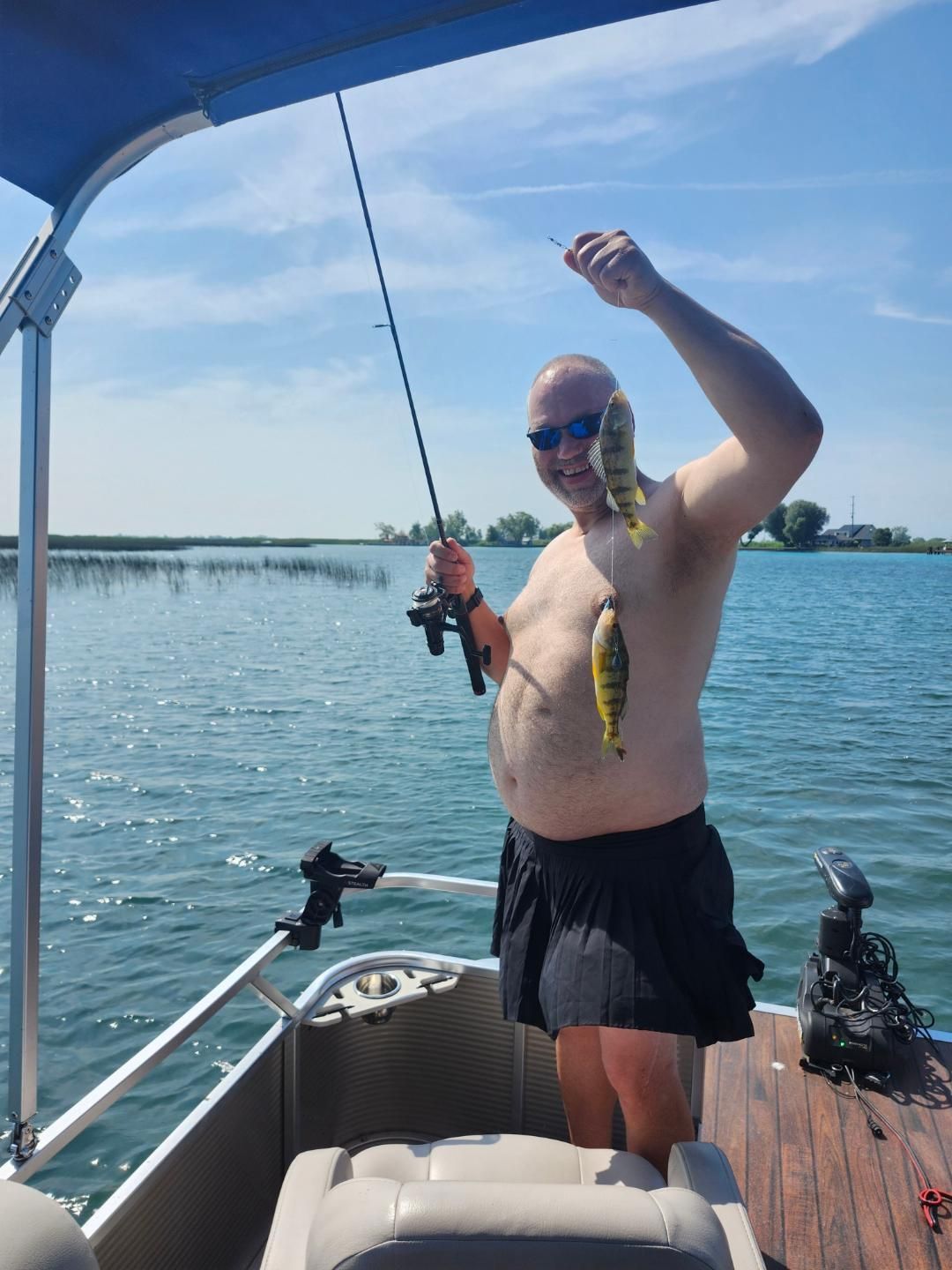 Man on a boat, holding up two yellow perch he caught, sunny day, water in background.