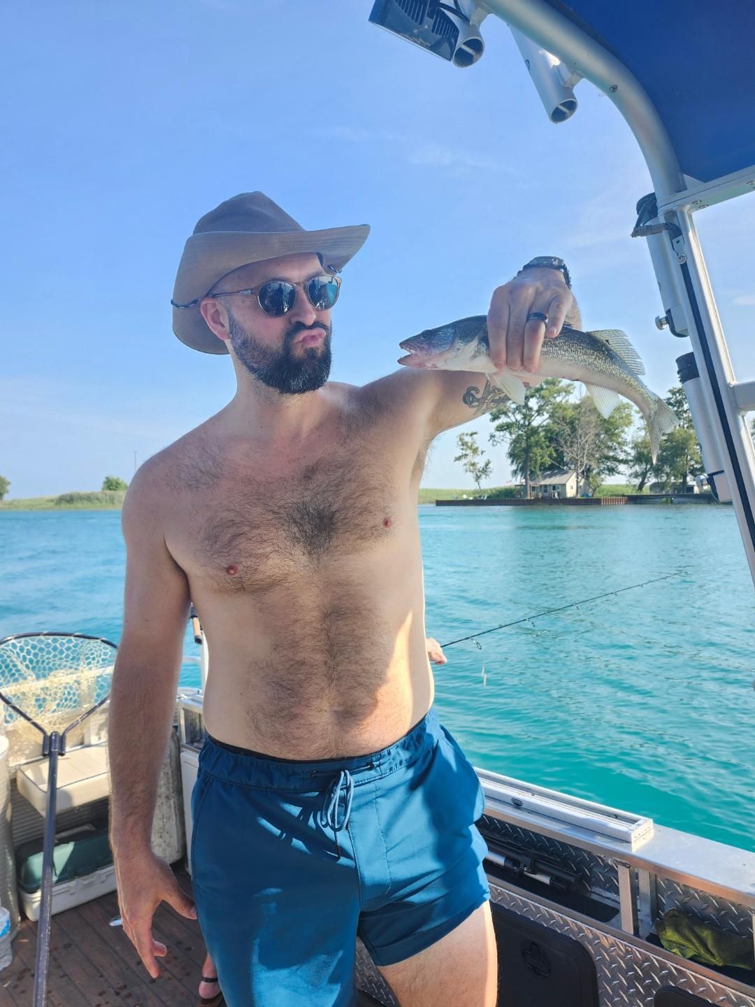 Man holding a fish on a boat; he wears sunglasses, a hat, and swim trunks; blue water in the background.