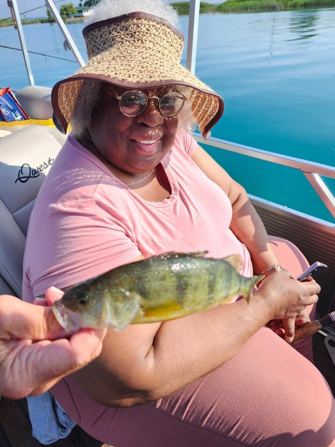 Woman on a boat holding a yellow perch she caught, wearing a hat and sunglasses, smiling.
