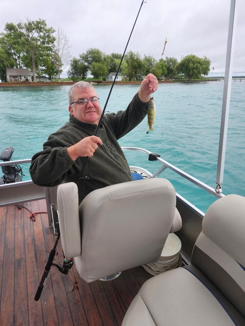 Man on a boat holds up a small fish. Turquoise water, trees, and cloudy sky.