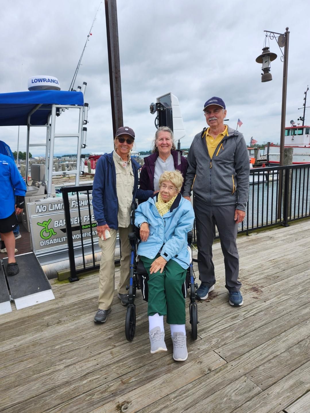 Four people on a pier, one in a wheelchair. Cloudy sky, boat in background.