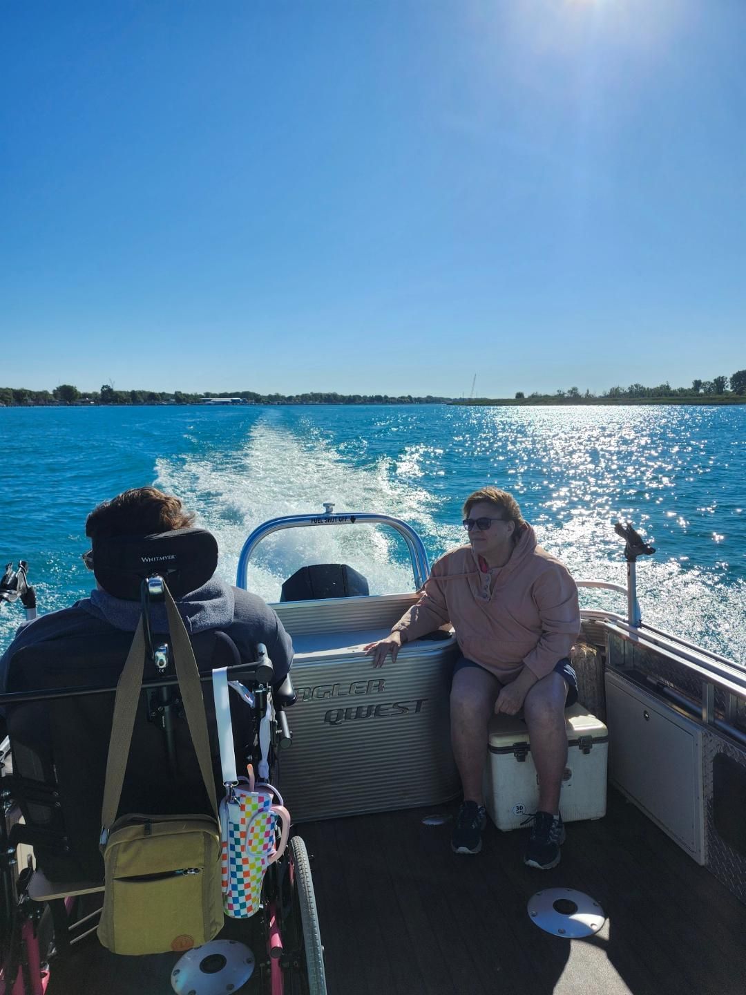 Person in wheelchair and another person on a boat on bright blue water; sunny day.