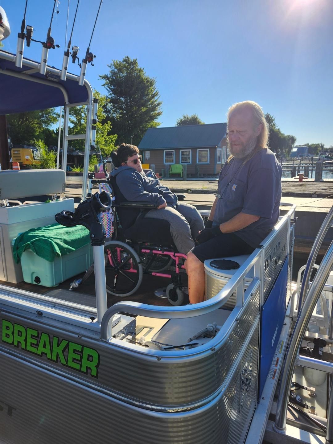 Man and person in wheelchair on a boat.  Person in wheelchair sits on the back with a man.  Fishing poles visible, sunny outdoors.