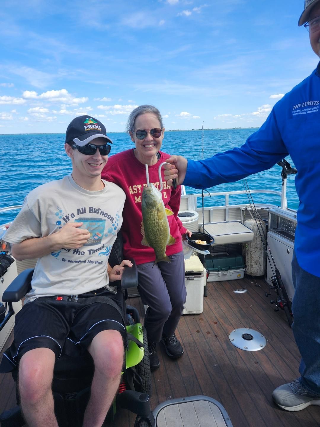 Person in wheelchair smiles on a boat, holding a fish. Another person holds up the fish. Blue water.