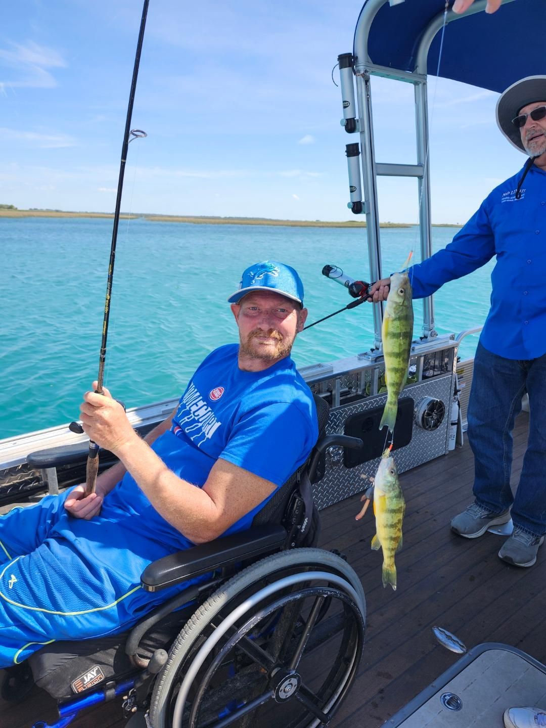 Man in wheelchair fishing on a boat, holding a rod. Another man displays two caught yellow perch. Blue water and sky.