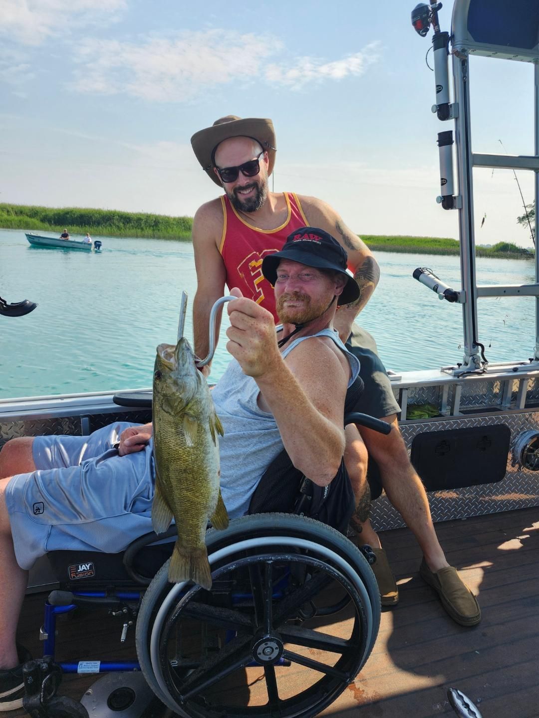 Man in wheelchair on a boat holding up a fish, another man behind him smiling. Water and sunny sky.