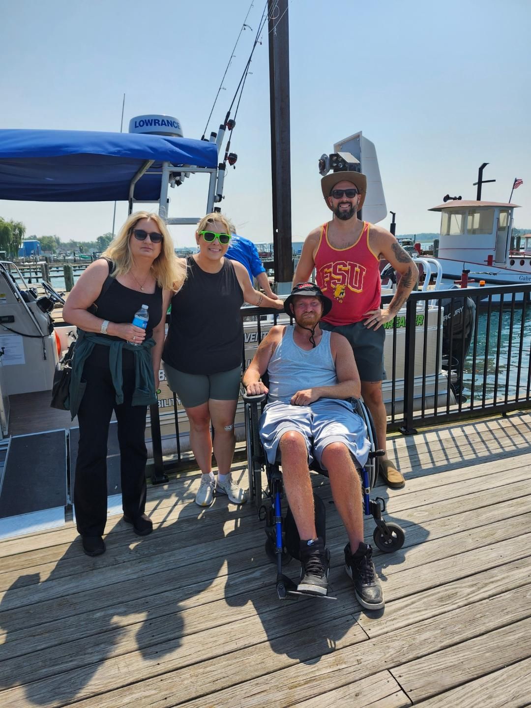Four people pose for photo on a dock near boats. One person sits in a wheelchair. Sunny day.
