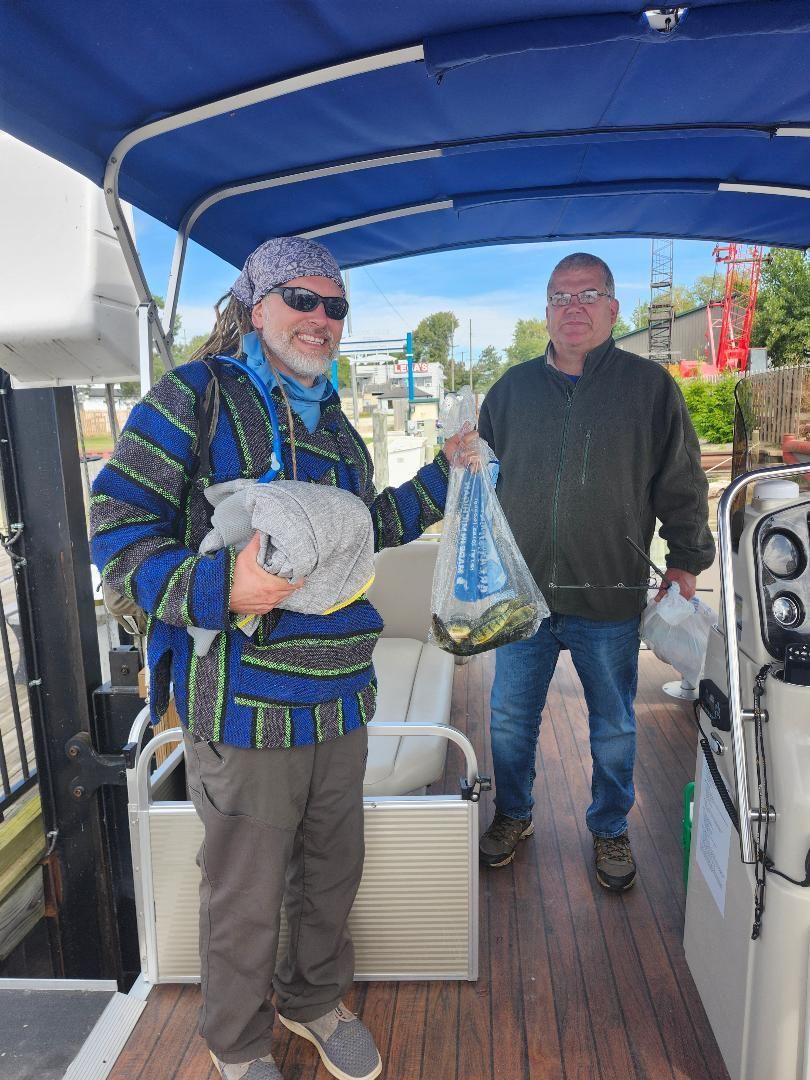 Two men on a boat holding bags. One wears a blue patterned sweater, the other a green shirt. Sunny day.