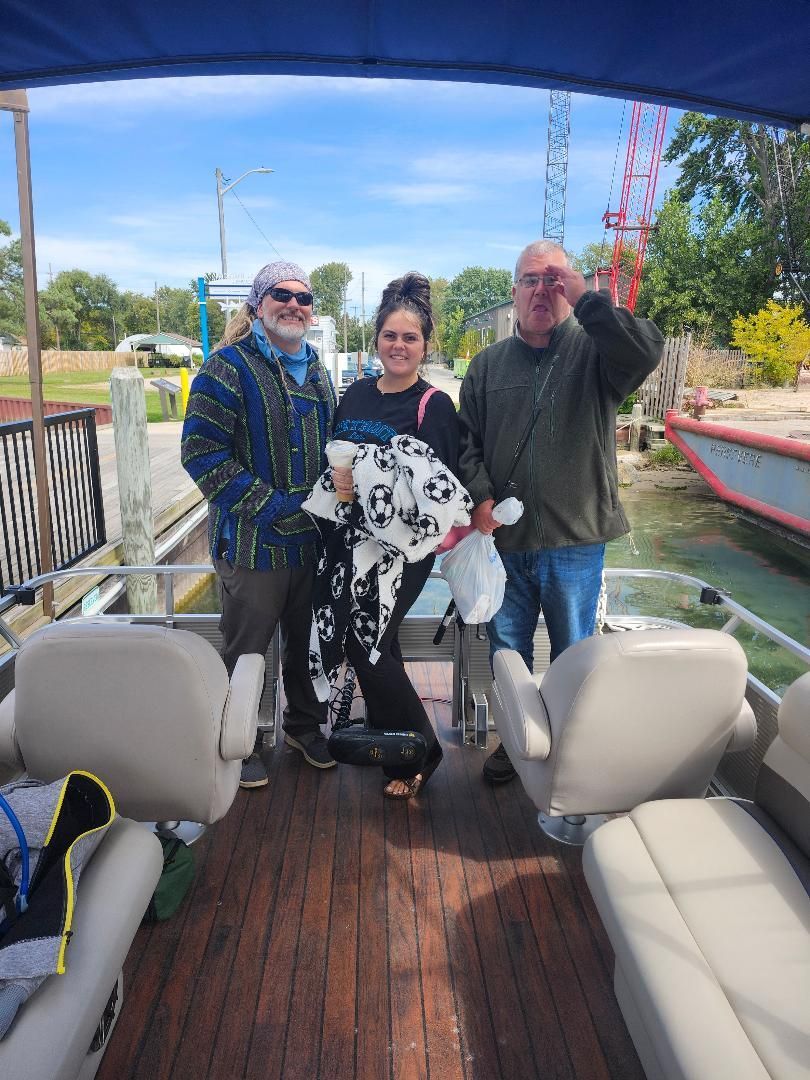 Three people on a boat near a dock. One waves, one holds a blanket, all smile. Outdoors on a sunny day.